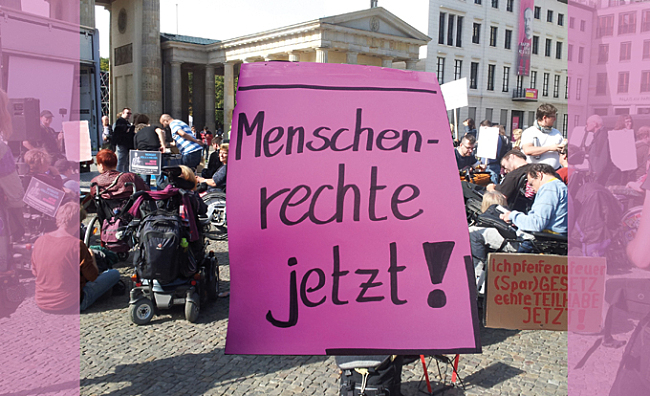 Foto: viele Menschen mit Plakaten auf einer Demo, viele sitzen im Rollstuhl, andere stehen oder sitzen auf dem Boden. Im Vordergrund übergroß ein in sehr großer Handschrift beschriebenes Plakat 