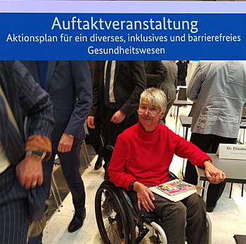 Sigrid Arnade sitz im Rollstuhl im Tagungsraum. Sie hat Informationsmaterial auf den Knien und lächelt skeptisch in die Kamera. An ihr vorbei gehen Menschen in Herrenanzügen, durch den Bildausschnitt sind keine Gesichter zu sehen.