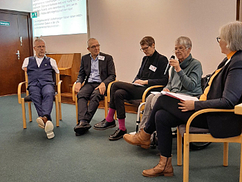 2 Frauen und 3 Männer sitzen in einem Halbkreis in einem Tagungsraum. Im Hintergrund eine Beamerprojektion mit Text sowie ein Klavier. Prof. Dr. Sigrid Arnade spricht in ein Mikrofon, das sie in der Hand hält.