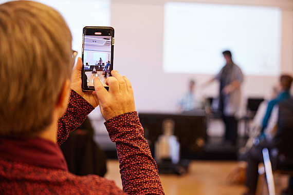 Fotos von der Welttagsveranstaltung: der Hinterkopf einer Frau, sie hält ein Handy hoch um ein Foto zu machen. Im Hintergrund verschwommen die Bühne