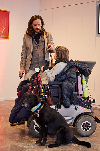 Austausch - Mark Wenzel (Musik) unterhält sich stehend mit einer Frau im E-Rollstuhl mit Assistenzhund, der neben ihr sitzt.