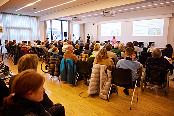 Der voll besetzte Tagungsraumes. Am linken Rand die Gebärdensprachdolmetscherin, an der Wand hinter der Bühne zwei Beamerprojektionen