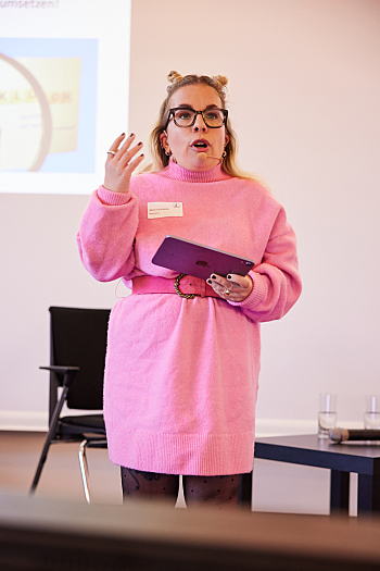 Begrüßung -Ninia LaGrande, Moderatorin durch die Tagung, in einem Strickkleid mit Gürtel in Pink und 2 Hörnchen aus Haar auf dem Kopf.