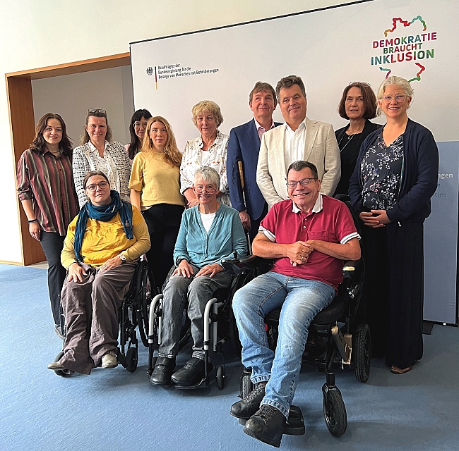 Auf dem Bild ist der Bundesbeauftragte für Menschen mit Behinderungen Jürgen Dusel zu sehen, umringt von Vertreter*innen des Deutschen Behindertenrats.